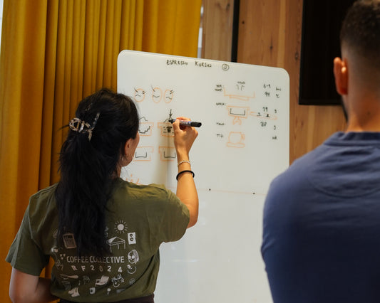 Person writing on a whiteboard with another person observing, against a yellow curtain.