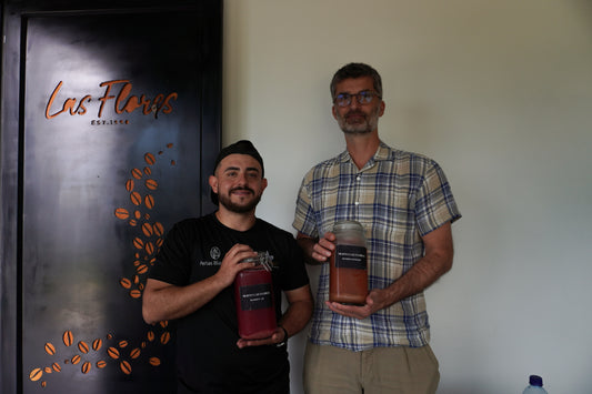 Two men holding jars in front of a 'Las Flores' branded door.