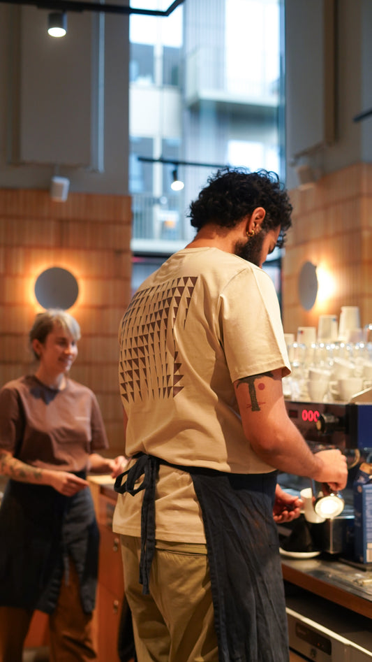 Person working behind a counter  in a cafe with another person in the background wearing Coffee Collective t-shirts